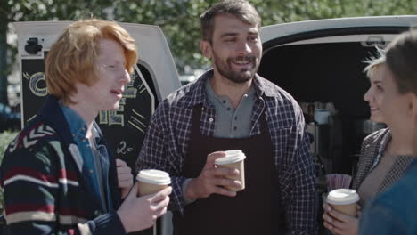 Young-Barista-With-Beard-Wearing-Apron-Standing-Before-His-Van-Drinking-Coffee-With-Young-People-And-Chatting