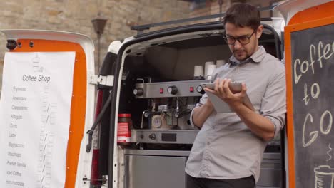 Male-Barista-Checking-Tablet-Leaned-Against-Mobile-Coffee-Van-Parked-In-Downtown-Street,