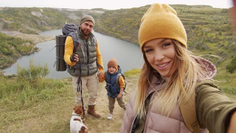 Pov-Shot-Of-Happy-Young-Woman-Holding-Camera-And-Taking-Selfie-Or-Filming-Herself-And-Her-Cheerful-Husband,-5-Year-Old-Son-And-Cute-Jack-Russell-Terrier-Dog-While-Hiking-Near-Quarry-Lake