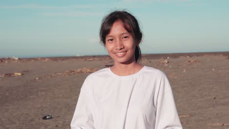 Medium-Closeup-Of-Young-Female-Indonesian-Teen-Collecting-Litter-From-Dirty-Polluted-Beach-Putting-It-In-Garbage-Bag-Then-Looking-At-Camera-Smiling