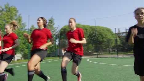 Team-Of-Young-Female-Soccer-Players-In-Uniform-Running-On-Field-Together-While-Training-Outdoors-On-Sunny-Summer-Day