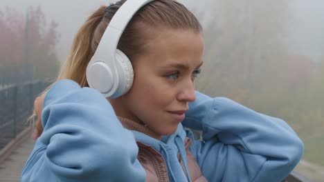 Handheld-Close-Up-Shot-Of-Beautiful-Young-Woman-Putting-Headphones-On-While-Preparing-For-Jog-Outdoors-On-Foggy-Autumn-Morning