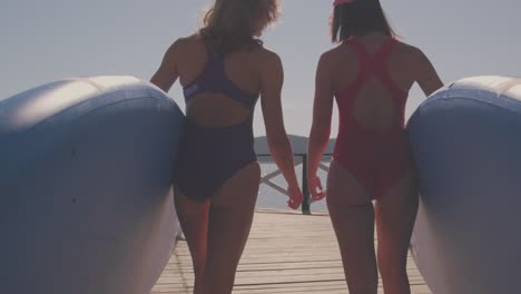 Medium-Back-View-Shot-Of-Two-Good-Looking-Girls-Holding-Paddle-Boards-In-Hands-Getting-Ready-For-Surfing