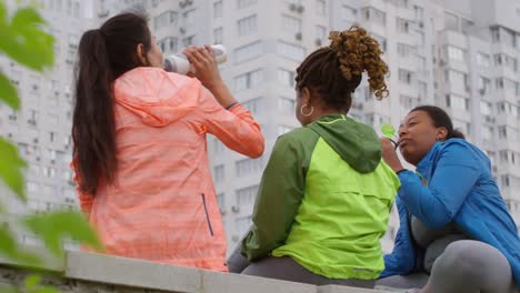 Rear-Shot-Of-Three-Female-Multiethnic-Friends-In-Bright-Neon-Sport-Jackets-Sitting-On-Stony-Parapet,-With-High-Rise-Apartment-Building-In-Background,-Drinking-Water-And-Taking-Selfies-On-Smartphone