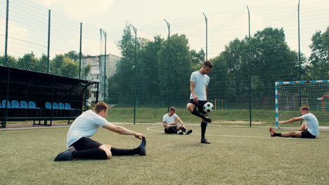 Young-Street-Football-Player-Training-Freestyle-Tricks-While-His-Team-Sitting-On-The-Pitch-Doing-Stretch-Exercises