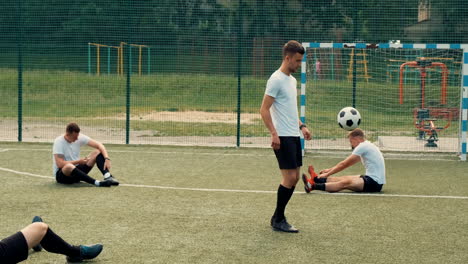 Young-Street-Football-Player-Training-Freestyle-Tricks-While-Two-Team-Players-Sitting-On-The-Pitch-Doing-Stretch-Exercises-And-Another-One-Using-Mobile-Phone-2