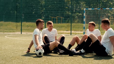Young-Street-Football-Players-Sitting-On-Pitch,-Resting-And-Talking-To-Each-Other-On-A-Sunny-Day