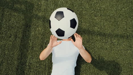 Top-View-Of-A-Happy-Young-Soccer-Player-Lying-On-Pitch,-Throwing-And-Catching-Ball-During-Training-Session
