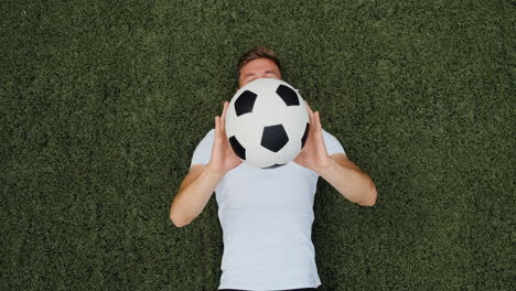 Top-View-Of-A-Happy-Young-Soccer-Player-Lying-On-Pitch,-Throwing-And-Catching-Ball-During-Training-Session-1