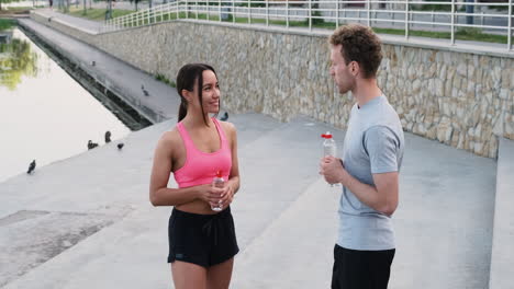 Happy-Sportive-Couple-Drinking-Water-And-Talking-To-Each-Other-While-Taking-A-Break-During-Their-Running-Session-In-The-City