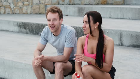 Beautiful-Sportive-Woman-Drinking-Water-And-Talking-With-Her-Boyfriend-While-They-Are-Sitting-On-Stairs-Taking-A-Break-During-Their-Running-Session-Outdoors