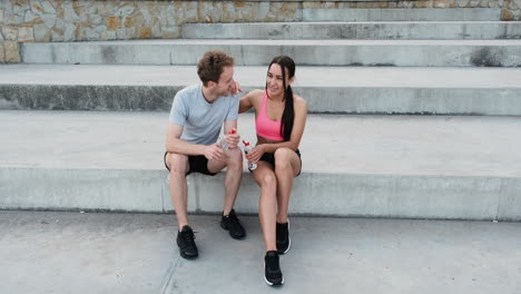 Happy-Sportive-Couple-Sitting-On-Stairs-Outdoors,-Holding-Water-Bottles-And-Talking-To-Each-Other-While-Taking-A-Break-During-Their-Running-Session-In-The-City