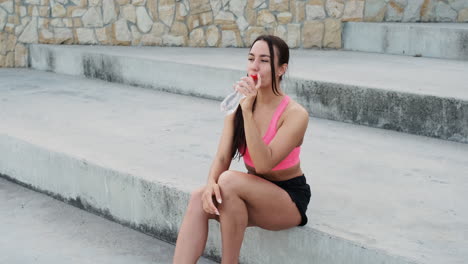 Beautiful-Sportive-Girl-Sitting-On-Stairs-And-Drinking-Water-After-Running-Training-Session-In-The-City