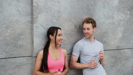 Happy-Sportive-Couple-Leaning-Against-A-Wall,-Holding-Drinking-Bottle-And-Taking-A-Break-During-Running-Session-In-The-City