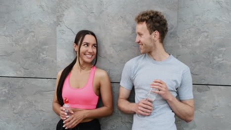 Happy-Sportive-Couple-Leaning-Against-A-Wall,-Holding-Water-Bottle-And-Talking-To-Each-Other,-While-Taking-A-Break-During-Running-Session-In-The-City