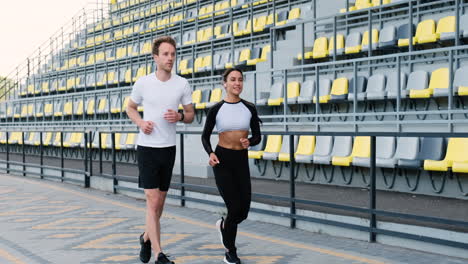 Happy-Runner-Girl-Wearing-Airpods-And-Sportive-Man-Checking-His-Smartwach-Running-Together-In-A-Stadium