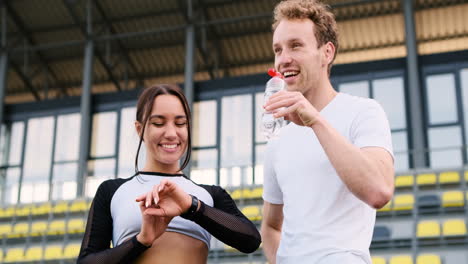Sportive-Young-Man-Checking-His-Girlfriend's-Smartwatch,-While-Having-A-Funny-Conversation-With-Her-And-Drinking-Water-In-The-Stadium