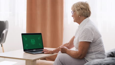 Seniorin-Sitzt-Auf-Sofa-Im-Wohnzimmer-Und-Spricht-über-Videoanruf-Auf-Modernem-Laptop-Mit-Grünem-Bildschirm