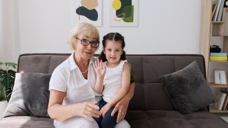 Happy-Grandmother-With-Her-Pretty-Little-Granddaughter-Sitting-On-Her-Knees-On-Sofa-Greeting-And-Talking-On-Video-Call