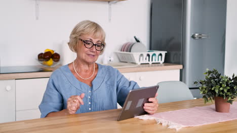 Feliz-Anciana-Sentada-A-La-Mesa-En-La-Cocina-Saludando-Y-Hablando-Por-Videollamada-Usando-Una-Tableta-Digital