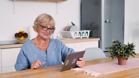 Glückliche-Seniorin,-Die-Am-Tisch-In-Der-Küche-Sitzt-Und-über-Einen-Videoanruf-Mit-Einem-Digitalen-Tablet-Spricht