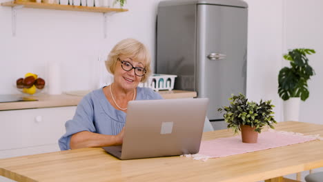 Glückliche-Seniorin,-Die-Am-Tisch-In-Der-Küche-Sitzt-Und-über-Videoanruf-Auf-Einem-Modernen-Laptop-Spricht