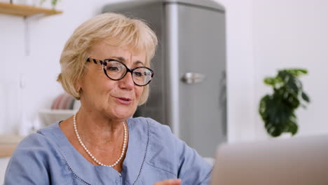 Porträt-Einer-Glücklichen-Seniorin,-Die-Am-Tisch-In-Der-Küche-Sitzt-Und-über-Einen-Videoanruf-Auf-Einem-Modernen-Laptop-Spricht