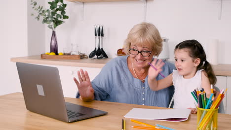 Kleines-Mädchen,-Das-Mit-Ihrer-Glücklichen-Großmutter-Am-Tisch-In-Der-Küche-Sitzt,-Während-Sie-Einen-Videoanruf-Auf-Einem-Modernen-Laptop-Hat