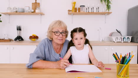 Grandmother-And-Granddaughter-Looking-At-Camera-During-A-Video-Call-While-Drawing-Together-Sitting-At-Table-In-Kitchen