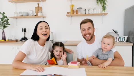 Happy-Young-Parents-Sitting-At-Table-In-Kitchen-Playing-With-Their-Two-Children-While-Looking-At-Camera-During-A-Video-Call