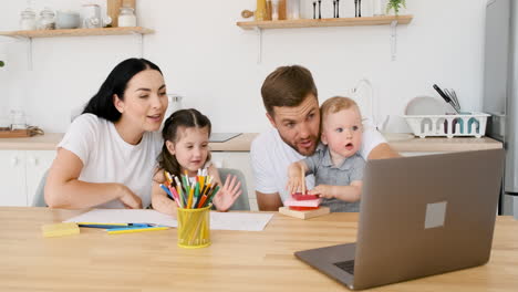 Glückliche-Eltern,-Die-Am-Tisch-In-Der-Küche-Sitzen-Und-Mit-Ihren-Beiden-Kindern-Spielen,-Während-Sie-Einen-Videoanruf-Auf-Einem-Modernen-Laptop-Führen