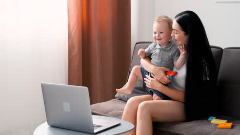 Beautiful-Mom-Sitting-On-Sofa-In-Living-Room-Holding-Her-Adorable-Little-Boy-While-Having-A-Video-Call-On-Moder-Laptop-At-Home
