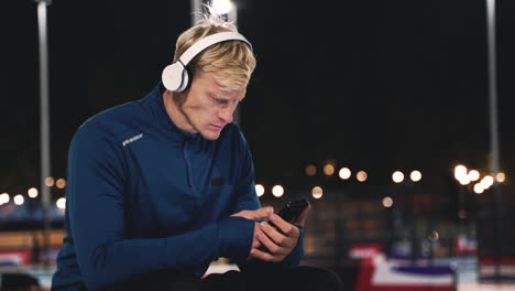 Sportive-Blond-Man-Sitting-At-Park-Listening-Music-With-Bluetooth-Headphones-And-Using-Mobile-Phone-While-Taking-A-Break-During-His-Training-Session-At-Night