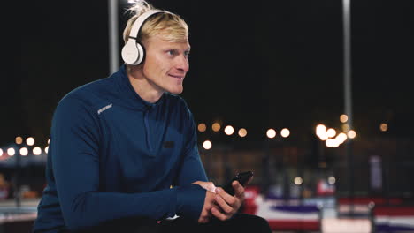 Smiling-Sportsman-Sitting-At-Park-Listening-Music-With-Bluetooth-Headphones,-Texting-On-His-Mobile-Phone-And-Looking-Around-Him-While-Taking-A-Break-During-His-Training-Session-At-Night
