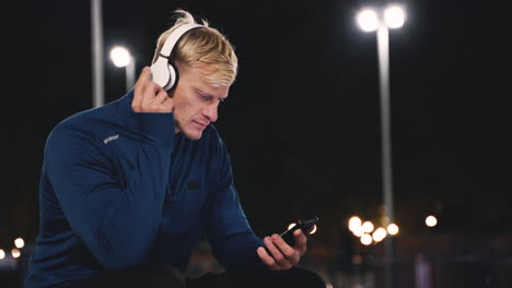 Smiling-Sportsman-Sitting-At-Park-Listening-Music-With-Bluetooth-Headphones-And-Texting-On-His-Mobile-Phone-While-Taking-A-Break-During-His-Training-Session-At-Night