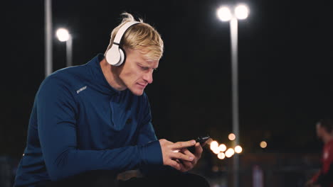 Sportive-Blond-Man-Sitting-At-Park-Listening-Music-With-Bluetooth-Headphones-And-Using-Mobile-Phone-While-Taking-A-Break-During-His-Training-Session-At-Night-1