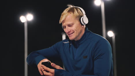 Happy-Sportsman-Sitting-At-Park-Listening-Music-With-Bluetooth-Headphones-And-Using-Mobile-Phone-While-Taking-A-Break-During-His-Training-Session-At-Night