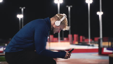 Sportive-Blond-Man-Sitting-At-Park-Listening-Music-With-Bluetooth-Headphones-And-Using-Mobile-Phone-While-Taking-A-Break-During-His-Training-Session-At-Night-2