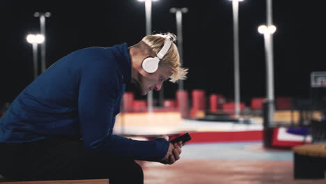 Smiling-Sportsman-Sitting-At-Park-Listening-Music-With-Bluetooth-Headphones,-Texting-On-His-Mobile-Phone-And-Looking-Around-Him-While-Taking-A-Break-During-His-Training-Session-At-Night-1