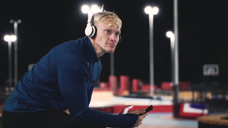Smiling-Sportsman-Sitting-At-Park-Listening-Music-With-Bluetooth-Headphones,-Texting-On-His-Mobile-Phone-And-Looking-Around-Him-While-Taking-A-Break-During-His-Training-Session-At-Night-2