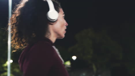 Sportive-Curly-Girl-Using-Mobile-Phone-To-Activate-Bluetooth-Headphones-And-Then-Start-To-Run-In-The-Park-At-Night