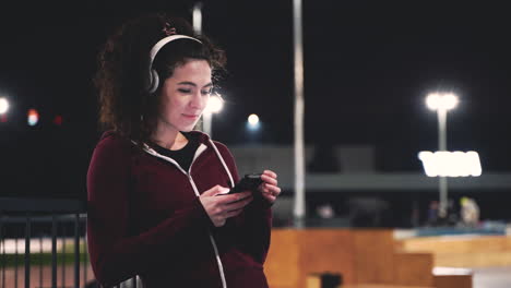 Smiling-Sportswoman-Listening-Music-With-Bluetooth-Headphones-And-Texting-On-Her-Mobile-Phone-While-Taking-A-Break-During-Her-Training-Session-At-Night-In-The-Park