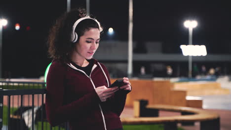 Smiling-Sportswoman-Listening-Music-With-Bluetooth-Headphones,-Texting-On-Her-Mobile-Phone-And-Looking-Around-While-Taking-A-Break-During-Her-Training-Session-At-Night-In-The-Park