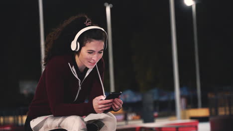 Smiling-Sportive-Woman-Sitting-At-Park-Listening-Music-With-Bluetooth-Headphones-And-Texting-On-Her-Mobile-Phone-While-Taking-A-Break-During-Her-Training-Session-At-Night