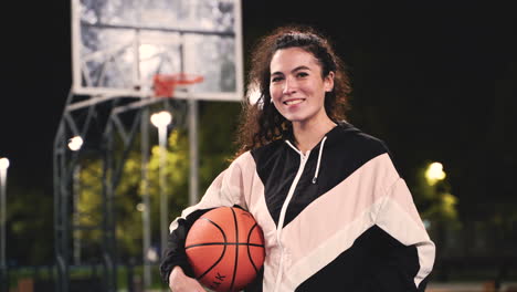 Porträt-Einer-Schönen-Basketballspielerin,-Die-Ball-Hält-Und-Nachts-Auf-Dem-Außenplatz-In-Die-Kamera-Lächelt