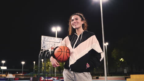 Schöne-Basketballspielerin,-Die-Ball-Hält-Und-Nachts-Auf-Dem-Außenplatz-In-Die-Kamera-Lächelt