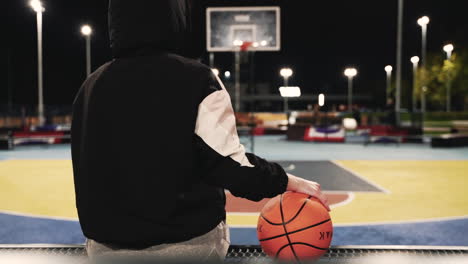 Back-View-Of-A-Female-Basketball-Player-In-Hoodie-Sitting-And-Holding-Ball-While-Taking-A-Break-After-Her-Training-Session-On-Outdoor-Court-At-Night