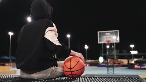 Back-View-Of-A-Female-Basketball-Player-In-Hoodie-Sitting-And-Holding-Ball-While-Taking-A-Break-After-Her-Training-Session-On-Outdoor-Court-At-Night-1