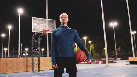 Confident-Basketball-Player-Holding-Ball-And-Looking-At-Camera-While-Standing-On-An-Outdoor-Court-At-Night