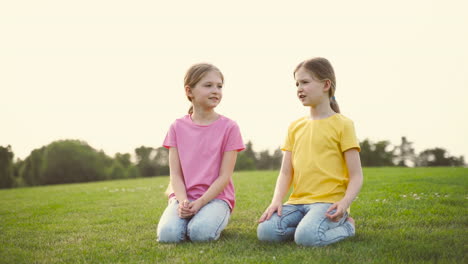 Two-Pretty-Little-Sisters-Talking-Together,-Looking-Around-And-Pointing-Something-While-Kneeling-On-Grass-At-Park